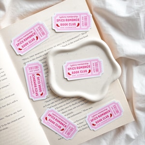 May include: Pink and white stickers with the text "Lifetime membership SPICY ROMANCE BOOK CLUB HEAT GUARANTEED" and a chili pepper graphic. The stickers are arranged on an open book and a white, wavy-edged dish.