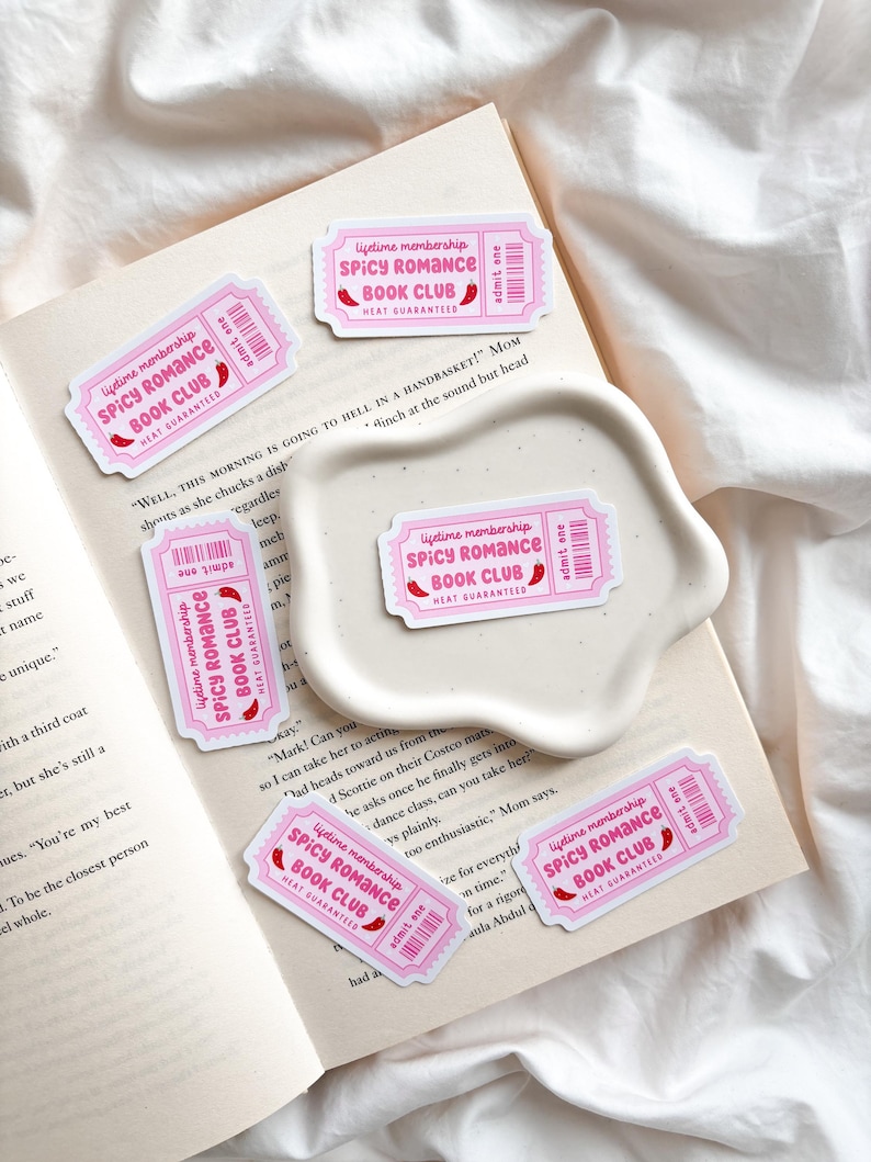 May include: Pink and white stickers with the text "Lifetime membership SPICY ROMANCE BOOK CLUB HEAT GUARANTEED" and a chili pepper graphic. The stickers are arranged on an open book and a white, wavy-edged dish.