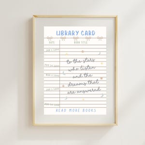 May include: A framed print featuring a vintage-style library card design. The top of the print reads "LIBRARY CARD" in blue script. The card includes lines for dates and book titles, with the text "to the stars who listen and the dreams that are answered."