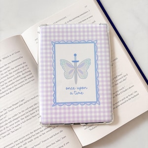 Könnte beinhalten: Eine hellviolett-weiß karierte Tablet-Hülle mit einem blau umrandeten Design. Das Design zeigt einen Schmetterling, ein Schwert und die Worte "once upon a time". Die Hülle liegt auf einem offenen Buch.