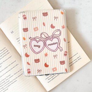 May include: A clear phone case with a cream and beige striped pattern. The case features a design with bears, bows, leaves, coffee cups, and the words "cosy reader" in a heart-shaped design. The case is resting on an open book.