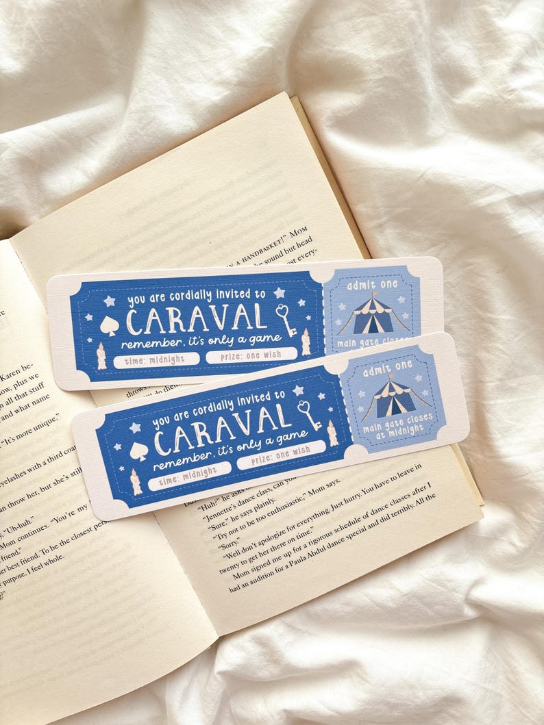 May include: Two blue Caraval-themed ticket-shaped bookmarks with white text and illustrations, including a key, stars, and a tent. The text reads "You are cordially invited to Caraval, remember, it's only a game." The bookmarks are placed on an open book.