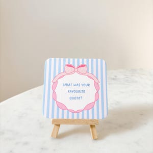May include: A small, square card with blue and white stripes, featuring a pink bow and a decorative frame. The text "WHAT WAS YOUR FAVOURITE QUOTE?" is inside the frame. The card is displayed on a miniature wooden easel.