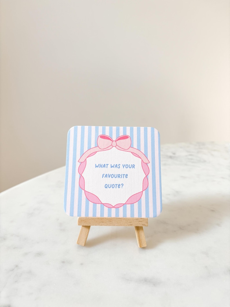May include: A small, square card with blue and white stripes, featuring a pink bow and a decorative frame. The text "WHAT WAS YOUR FAVOURITE QUOTE?" is inside the frame. The card is displayed on a miniature wooden easel.