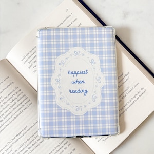 Peut inclure: Une coque de Kindle à carreaux bleu clair et blanc avec l'inscription "happiest when reading" dans un cadre décoratif. L'étui est posé sur un livre ouvert.