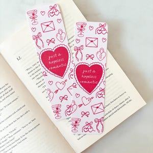 May include: Two white bookmarks with red illustrations of hearts, bows, champagne glasses, and envelopes. Each bookmark has a red heart with the text "just a hopeless romantic". The bookmarks are placed on an open book.