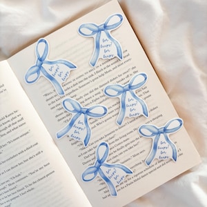 Könnte beinhalten: Sechs hellblaue Aufkleber in Schleifenform mit dem Text "for keeps?" sind auf den offenen Seiten eines Buches angeordnet. Die Aufkleber haben einen Aquarelleffekt, und die Seiten des Buches sind mit gedrucktem Text gefüllt.