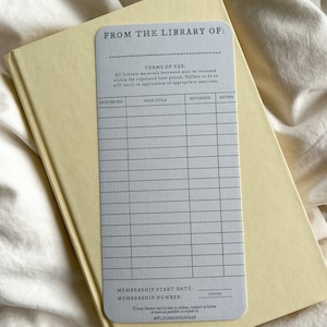 K&ouml;nnte beinhalten: Eine hellgraue Bibliothekskarte mit dem Text "FROM THE LIBRARY OF:" und einem Raster f&uuml;r Buchdetails, die auf einem hellgelben Buch liegt. Die Karte enth&auml;lt Felder f&uuml;r Ausgabedatum, Buchtitel, zur&uuml;ckgegeben und Bewertung. Der Text "@FLEURSONSUNDAY" befindet sich unten.