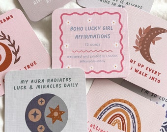 Könnte beinhalten: Eine Sammlung quadratischer Affirmationskarten mit verschiedenen Designs und Texten. Die Karten zeigen Illustrationen von Sonne, Mond, Regenbogen und Planeten. Die zentrale Karte lautet "BOHO LUCKY GIRL AFFIRMATIONS" und "12 Karten". Die Karten sind rosa und weiß.