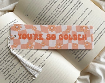 Puede incluir: Un marcapáginas con un patrón a cuadros melocotón y blanco y gráficos de margaritas sonrientes. El texto "YOU'RE SO GOLDEN" está impreso en naranja. Una borla blanca cuelga de la parte superior. El marcapáginas descansa sobre un libro abierto.