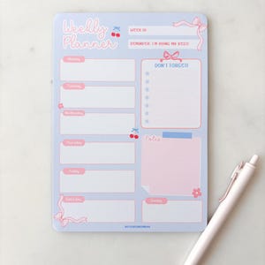 Op de afbeelding: Een lichtblauw weekplanner-notitieblok met roze accenten, voorzien van de tekst Weekly Planner, Don't Forget en Notes. Het ontwerp bevat illustraties van kersen, strikken en bloemen, met een herinneringssectie met de tekst I'm doing my best.