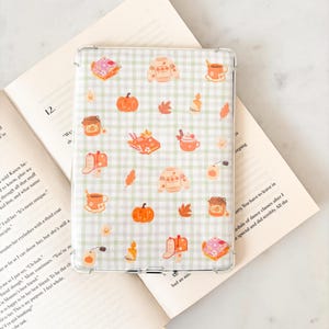 May include: Transparent phone case featuring a fall-themed design. The pattern includes pumpkins, sweaters, leaves, and mugs on a green and white checkered background. The case is on an open book.
