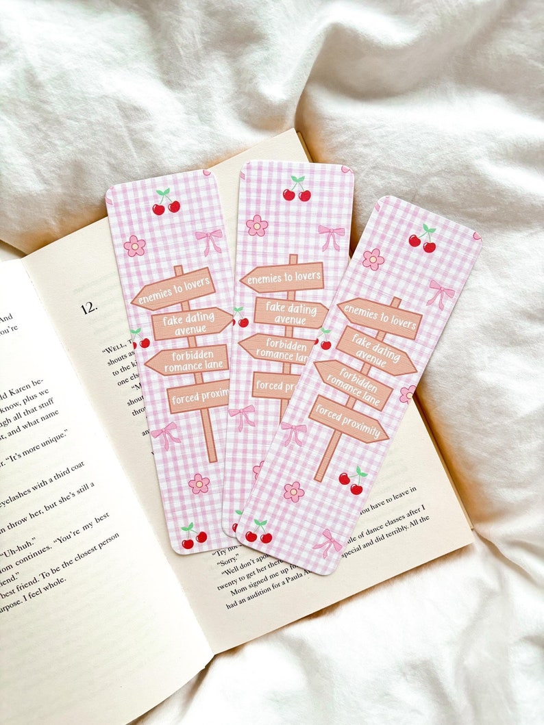 Puede incluir: Tres marcap&aacute;ginas rosas de cuadros vichy con detalles de cerezas y flores. Cada marcap&aacute;ginas presenta un dise&ntilde;o de se&ntilde;al de tr&aacute;fico con frases como "enemies to lovers" y "forced proximity". Los marcap&aacute;ginas est&aacute;n sobre un libro abierto.