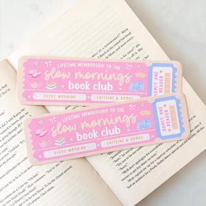 Peut inclure: Deux cartes de membre roses du "Slow Mornings Book Club" avec des motifs de billets, avec du texte et des illustrations de tasses à café, de livres et de pantoufles. Les cartes sont placées sur un livre ouvert.