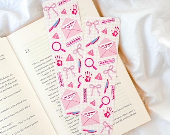 Op de afbeelding: Twee roze en wit gekleurde bladwijzers met misdaadthema-illustraties, waaronder een mes, vergrootglas, handafdruk, strik en envelop met de tekst "THE HUSBAND DID IT!" en "WARNING". De bladwijzers liggen op een open boek.
