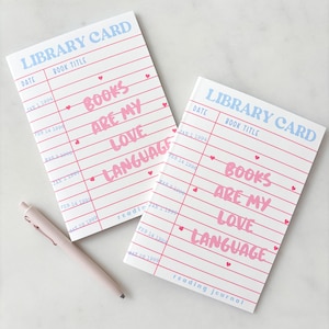 May include: Two rectangular reading journals with a white cover featuring a blue library card header, pink horizontal lines, and the pink text "BOOKS ARE MY LOVE LANGUAGE" with small hearts. A light pink pen is positioned next to the journals.