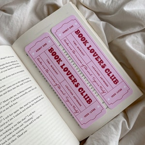 May include: Two pink "Book Lovers Club" ticket-shaped bookmarks with red text, resting on the open pages of a book. The bookmarks feature the text "Books are the mirrors of the soul - Virginia Woolf."