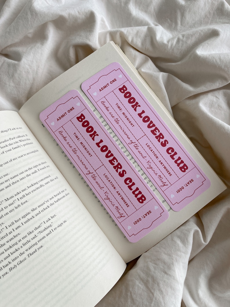 May include: Two pink "Book Lovers Club" ticket-shaped bookmarks with red text, resting on the open pages of a book. The bookmarks feature the text "Books are the mirrors of the soul - Virginia Woolf."