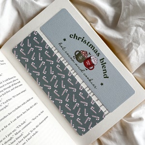 Op de afbeelding: Twee bladwijzers liggen op een open boek. Eén is lichtblauw met de tekst "christmas blend hot cocoa and cookies" en illustraties van mokken. De andere is donkergrijs met een zuurstokpatroon.