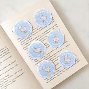 Op de afbeelding: Een verzameling achthoekige stickers met een lichtblauwe rand en de tekst "silly goose on the loose". Elke sticker toont een cartoon gans met een roze lichaam en een blauwe sjaal. De stickers liggen op een open boek.