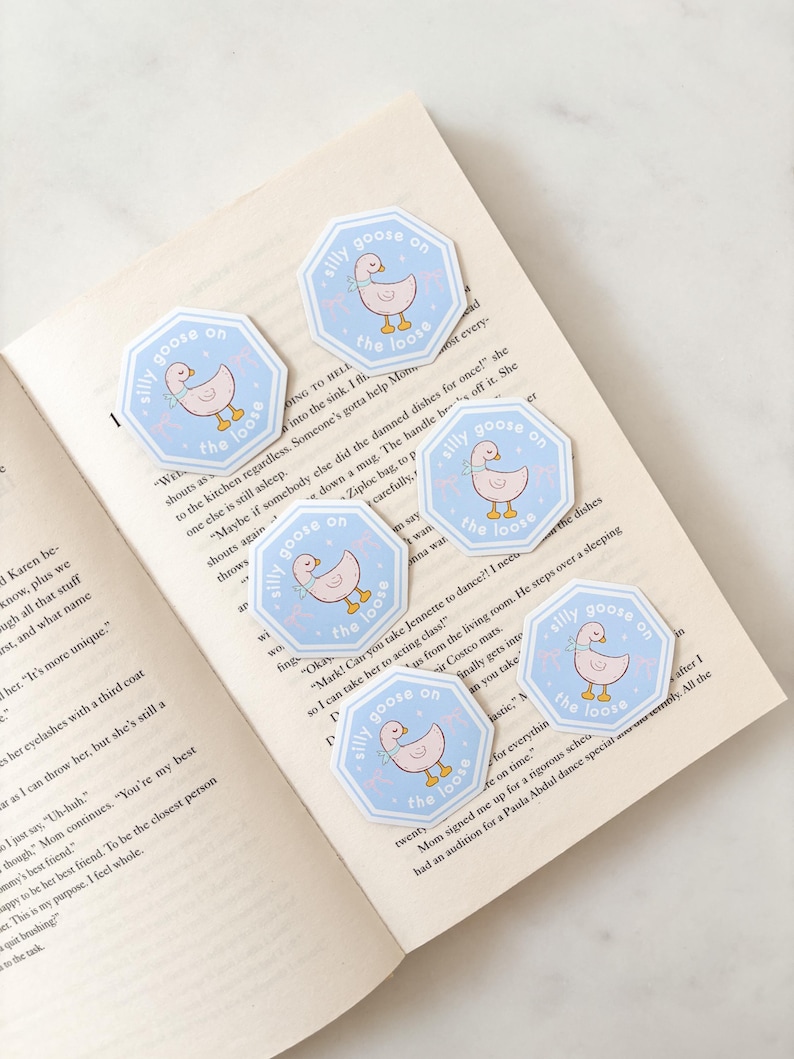 Op de afbeelding: Een verzameling achthoekige stickers met een lichtblauwe rand en de tekst "silly goose on the loose". Elke sticker toont een cartoon gans met een roze lichaam en een blauwe sjaal. De stickers liggen op een open boek.
