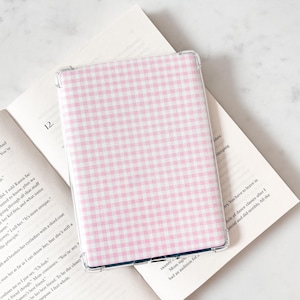 Może przedstawiać: Przezroczyste etui ochronne na czytnik e-booków, z różowo-białym wzorem w kratkę. Etui spoczywa na otwartej książce z nadrukowanym tekstem. Etui na czytnik dodaje stylu i zapewnia ochronę.