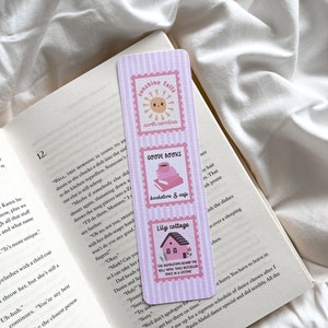 May include: A pink and white striped bookmark with three rectangular designs. The top design features a smiling sun and the text "sunshine falls north carolina." The middle design says "GOODE BOOKS bookstore & cafe." The bottom design shows a house and says "Lily cottage."