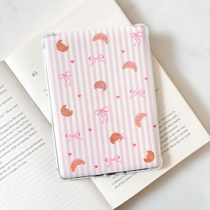 May include: A clear tablet case featuring a pink and white striped design. The case is adorned with illustrations of croissants, pink bows, and small pink hearts. The case is placed on an open book.