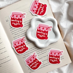 以下が含まれることがあります： 「reading is my jam」の文字が入った、赤いとピンク色のジャム瓶の形をしたステッカーのコレクション。各ステッカーにはイチゴのデザインがあり、開いた本の上に配置されています。