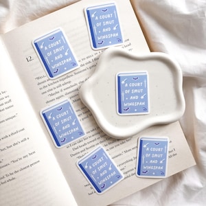 Op de afbeelding: Een verzameling lichtblauwe stickers met de tekst "A COURT OF SMUT AND WINGSPAN" in het wit. De stickers hebben een boekontwerp met vleermuis- en zwaardillustraties. Ze zijn gerangschikt op een open boek en een wit schaaltje.