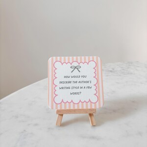 May include: A small, decorative sign on a wooden easel. The sign has a pink and white striped background with a scalloped edge and a bow graphic. Text on the sign reads, "HOW WOULD YOU DESCRIBE THE AUTHOR'S WRITING STYLE IN A FEW WORDS?"