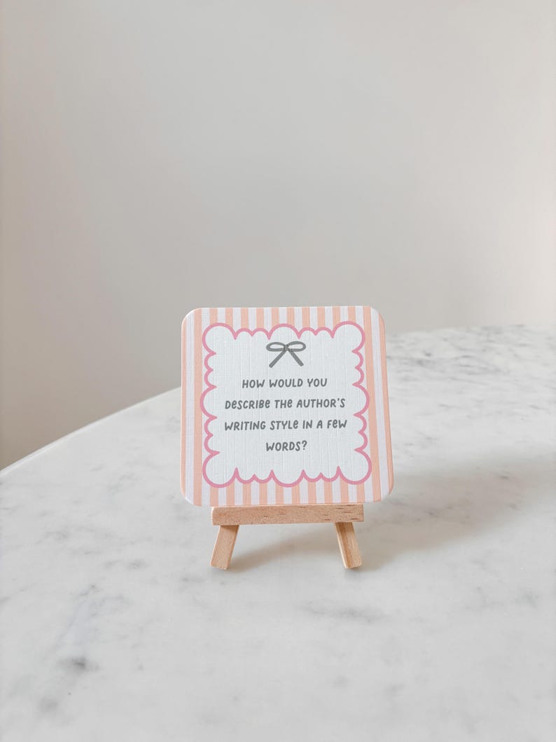 May include: A small, decorative sign on a wooden easel. The sign has a pink and white striped background with a scalloped edge and a bow graphic. Text on the sign reads, "HOW WOULD YOU DESCRIBE THE AUTHOR'S WRITING STYLE IN A FEW WORDS?"