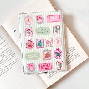 Könnte beinhalten: Transparente Tablet-Hülle mit weihnachtlichen Aufklebern. Die Aufkleber zeigen Lebkuchenmänner, Weihnachtsmänner, Schneemänner, Schleifen und Zuckerstangen. Auf den Aufklebern steht "Festive Book Club", "Admit One Sleigh Ride" und "Winter Book Club cozy vibes only".