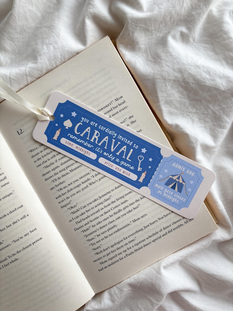 May include: A blue and white bookmark with the text "CARAVAL" and "Admit One" on it, placed in an open book. The bookmark has a ribbon attached. The text on the bookmark reads "you are cordially invited to CARAVAL, remember, it's only a game, time: midnight, prize: one wish."