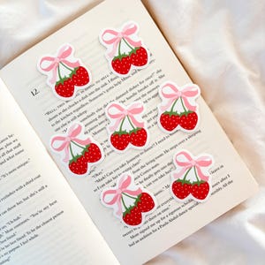 May include: A collection of strawberry stickers, each with a pink bow, are placed on the open pages of a book. The stickers depict two red strawberries with green stems and white outlines. The book's pages are cream-coloured.
