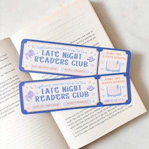May include: Two blue and white Late Night Readers Club bookmarks with text and illustrations, placed on an open book. The bookmarks feature phrases like "Just one more page" and "books + blankets."