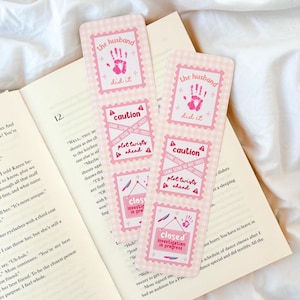 May include: Two pink and white gingham bookmarks with the text "the husband did it" and "caution crime scene do not cross plot twists ahead". The bookmarks also say "closed investigation in progress". The bookmarks are on top of an open book.