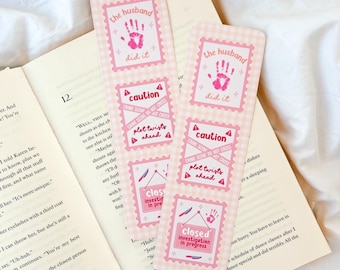 Op de afbeelding: Twee roze en witte gingham bladwijzers met de tekst "the husband did it" en "caution crime scene do not cross plot twists ahead". De bladwijzers zeggen ook "closed investigation in progress". De bladwijzers liggen op een open boek.