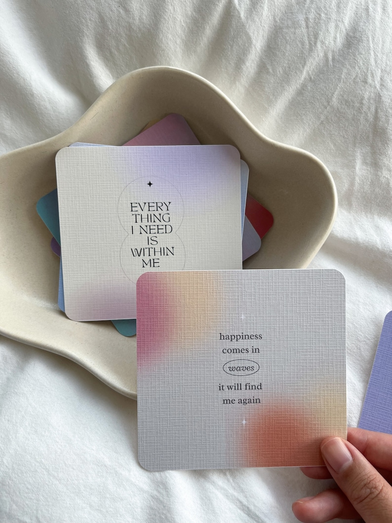 Puede incluir: Un mont&oacute;n de tarjetas cuadradas con citas inspiradoras y un dise&ntilde;o de gradiente suave y colorido. Una tarjeta dice "EVERYTHING I NEED IS WITHIN ME." Otra dice "happiness comes in waves it will find me again."