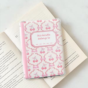 Può includere: Una custodia trasparente per Kindle con un motivo a ciliegie e fiocchi rosa. La custodia ha un dorso rosa e un'etichetta con la scritta "this kindle belongs to". Appoggiata su un libro aperto.