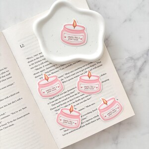 May include: A white, star-shaped dish holds a pink candle sticker with the text "smells like a plot twist." Several similar stickers are scattered on an open book. The candle design includes a flame and a heart.