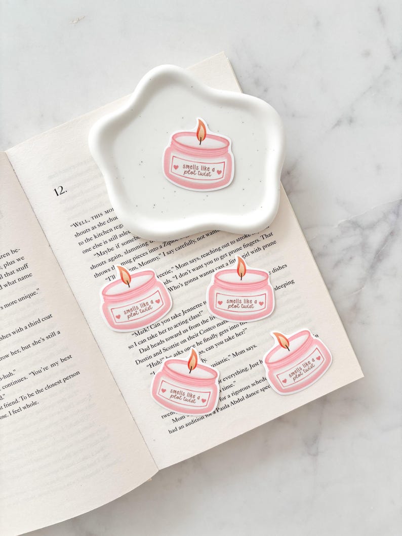May include: A white, star-shaped dish holds a pink candle sticker with the text "smells like a plot twist." Several similar stickers are scattered on an open book. The candle design includes a flame and a heart.