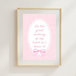 May include: A framed print with a pink background and a white oval design. The oval contains the text "it's the great undoing of my heart as I know it" in a script font. A pink bow is at the bottom of the oval.