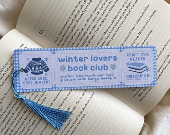 May include: A blue and white bookmark with the text "winter lovers book club" and a sweater graphic. It has a blue tassel and is placed on an open book. The bookmark reads "winter was made for hot cocoa and large books."
