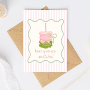 May include: A greeting card with a pink and white striped background. The card features a matcha drink illustration with the text "love you so matcha!". The card is inside a brown envelope.