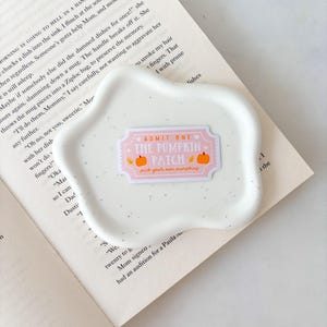May include: A white, speckled, wavy-edged ceramic dish with a pink and orange sticker that reads "ADMIT ONE THE PUMPKIN PATCH pick your own pumpkins." The dish rests on an open book.