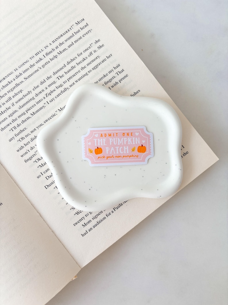 May include: A white, speckled, wavy-edged ceramic dish with a pink and orange sticker that reads "ADMIT ONE THE PUMPKIN PATCH pick your own pumpkins." The dish rests on an open book.