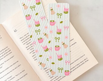 May include: Two decorative bookmarks with a light green and white striped background. The bookmarks feature a repeating pattern of pink teacups, matcha whisks, spoons, bows, and flowers. The bookmarks are resting on the open pages of a book.