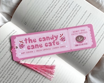 May include: Pink bookmark with the text "the candy cane cafe" and a tassel. The bookmark has a ticket design with the words "Hot Cocoa, Fresh Cookies, Candy Canes". The bookmark is placed on an open book.