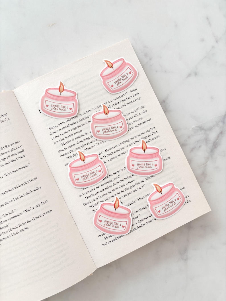 May include: A collection of pink candle stickers with the text "smells like a plot twist" and a small heart. The stickers are placed on the open pages of a book. The candles have a flame.